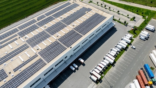 Aerial view of a large warehouse with solar panels on the roof and delivery trucks parked at the loading bay