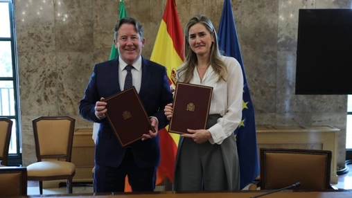 Two officials hold signed documents during a formal ceremony with Spanish and EU flags behind