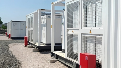 White containerised battery storage enclosures with wire mesh doors and red fire suppression boxes on gravel